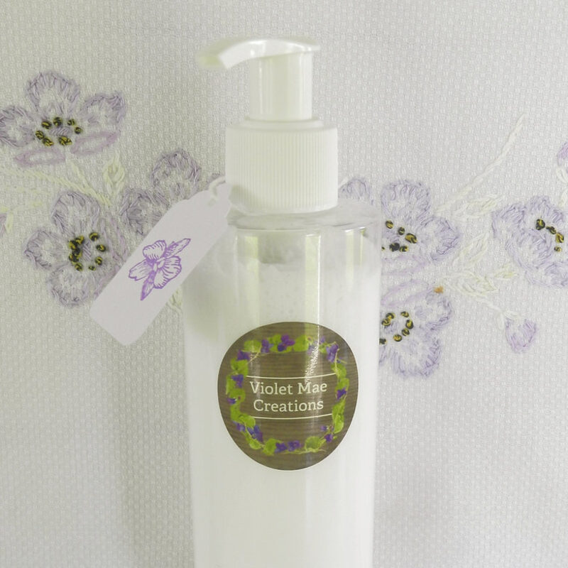 An 8 oz pump bottle of Violet Mae lotion with a hand-tied violet gift tag, against embroidered linen.