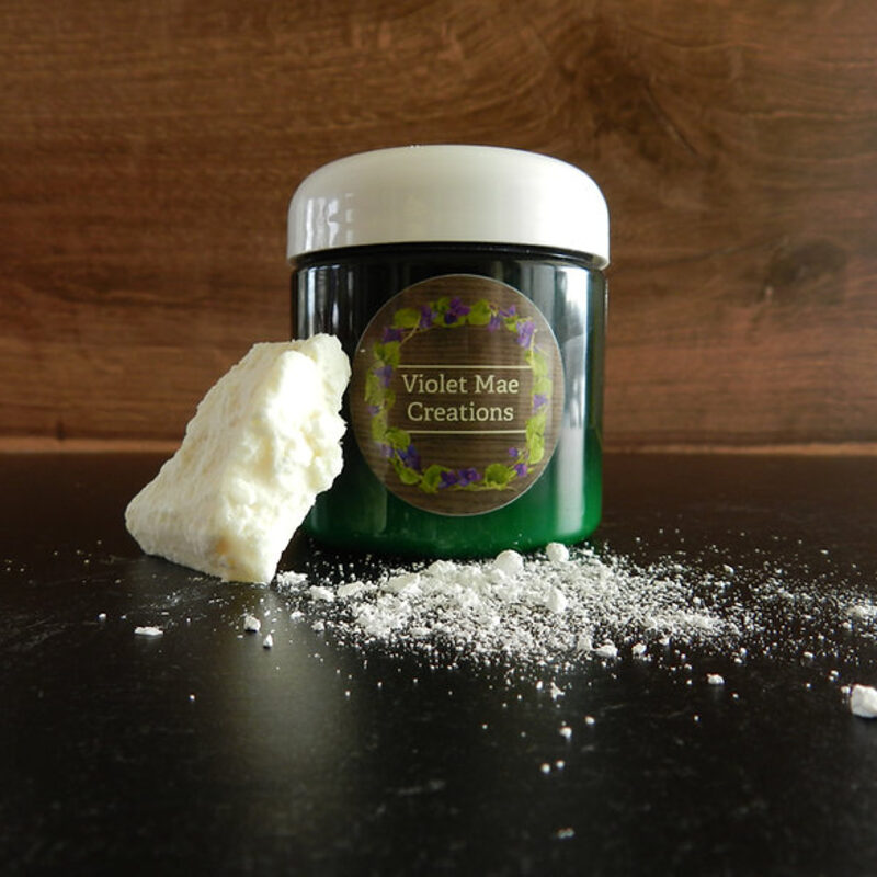 A 4 oz green glass jar of restorative cream beside a broken piece of raw shea butter.